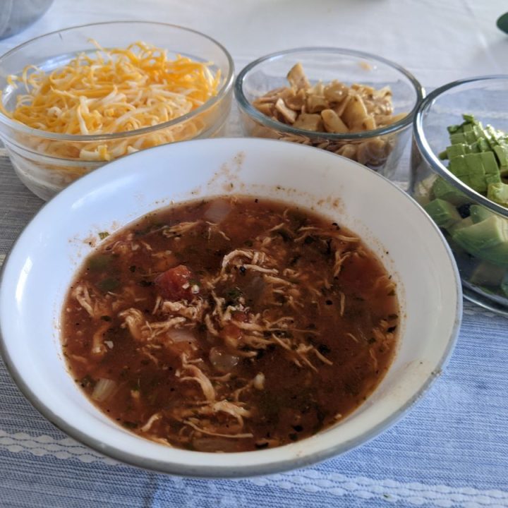 bowl of chicken tortilla soup, bowl with shredded cheese, bowl with tortilla strips, and bowl with avocado on a table