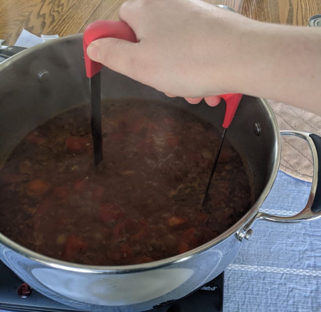 A hand using a potato masher to break apart tomatoes in chili