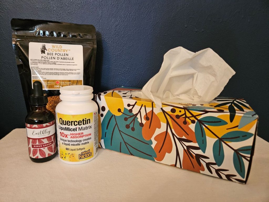 A bag of bee pollen, a dropper bottle of sinus saver, a bottle of quercetin and a box of tissue on a night stand.