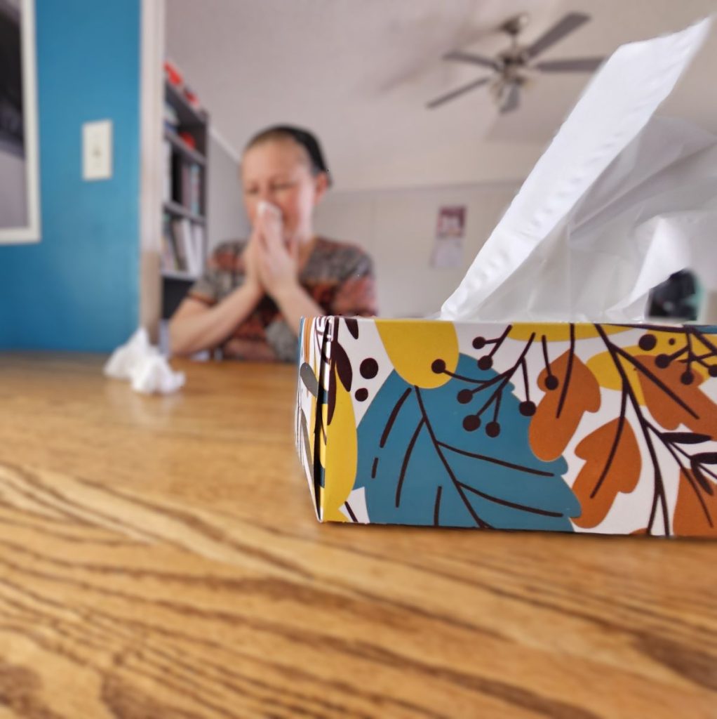 A Box of tissues on a table with a blurred back round of a woman blowing her nose